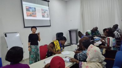 Instructor standing at the front of a classroom giving a presentation to a diverse group seated around tables with a projected slide behind her