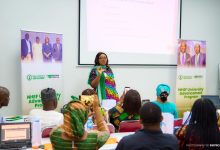 Presenter in a colorful outfit speaks to an audience at the front of a conference room with banners on both sides for the NHEF University Advancement Program, and a projected slide above her.
