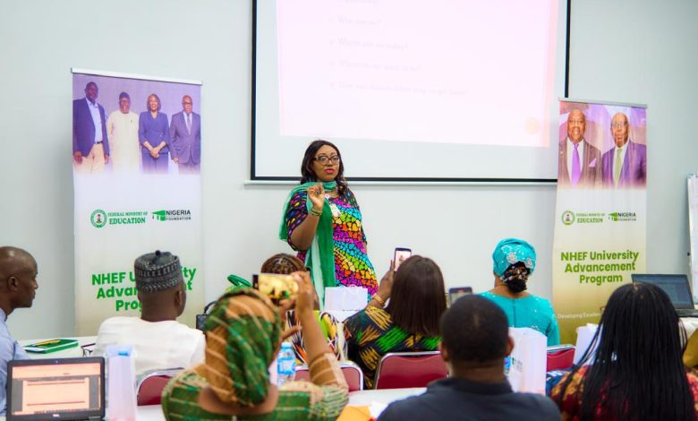 FGs university advancement programme - The Times of Abuja Presenter in a colorful outfit speaks to an audience at the front of a conference room with banners on both sides for the NHEF University Advancement Program, and a projected slide above her.