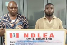 Two men stand indoors, each holding a framed NDLEA MMIA Command board with handwritten details.