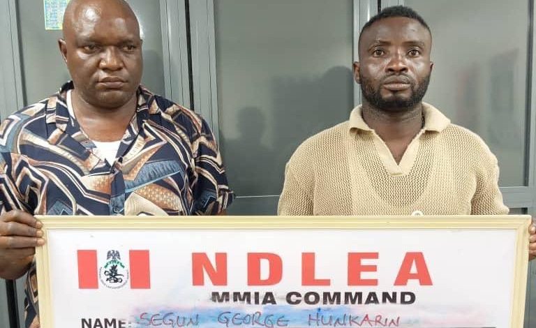 Two men stand indoors, each holding a framed NDLEA MMIA Command board with handwritten details.