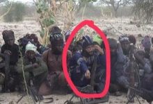 Group of armed men sitting on sandy ground in a dry, desert landscape; one man in a blue shirt at center raises his hand, circled in red, with rifles nearby.