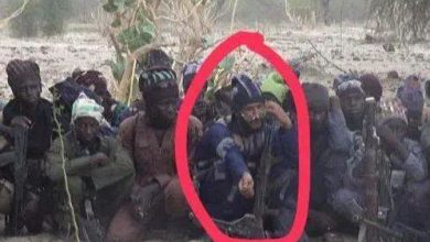 Group of armed men sitting on sandy ground in a dry, desert landscape; one man in a blue shirt at center raises his hand, circled in red, with rifles nearby.