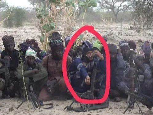 Group of armed men sitting on sandy ground in a dry, desert landscape; one man in a blue shirt at center raises his hand, circled in red, with rifles nearby.