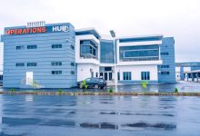 Blue modern office building labeled 'OPERATIONS HUB' with a parked car in front on a wet road, overcast sky nearby.