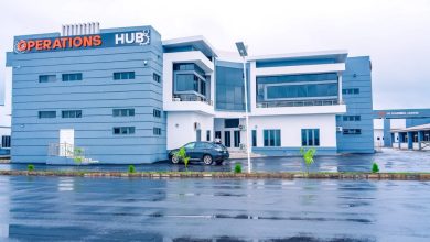 Blue modern office building labeled 'OPERATIONS HUB' with a parked car in front on a wet road, overcast sky nearby.