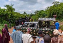 Military personnel in camouflage with armed trucks on a dirt rural road, as civilians gather nearby.