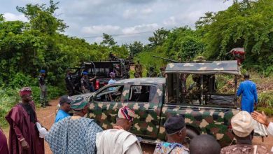 Military personnel in camouflage with armed trucks on a dirt rural road, as civilians gather nearby.
