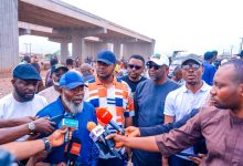 Bearded man in a blue cap speaks to reporters at a construction site under an overpass, with journalists and colorful microphones surrounding him.