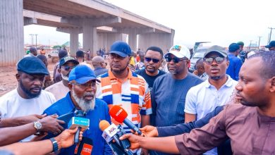 Bearded man in a blue cap speaks to reporters at a construction site under an overpass, with journalists and colorful microphones surrounding him.