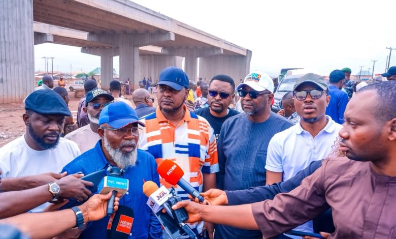 Bearded man in a blue cap speaks to reporters at a construction site under an overpass, with journalists and colorful microphones surrounding him.