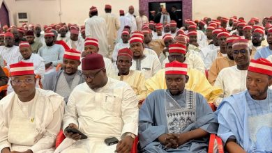 Large group of men in traditional white, cream, blue outfits and red caps seated at an indoor formal event.