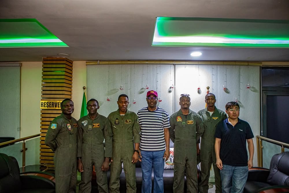 Eight men stand in a row indoors; six wear green flight-suits with patches, two civilians in casual clothing flank them, green neon lighting visible overhead, a 'RESERVE' sign is on a wooden pillar to the left.