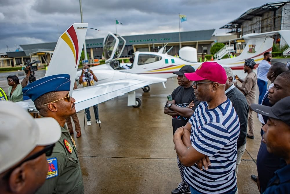 Group of men, including a man in a striped shirt and red cap, talking with a pilot in a blue hat beside a small white airplane on the tarmac.