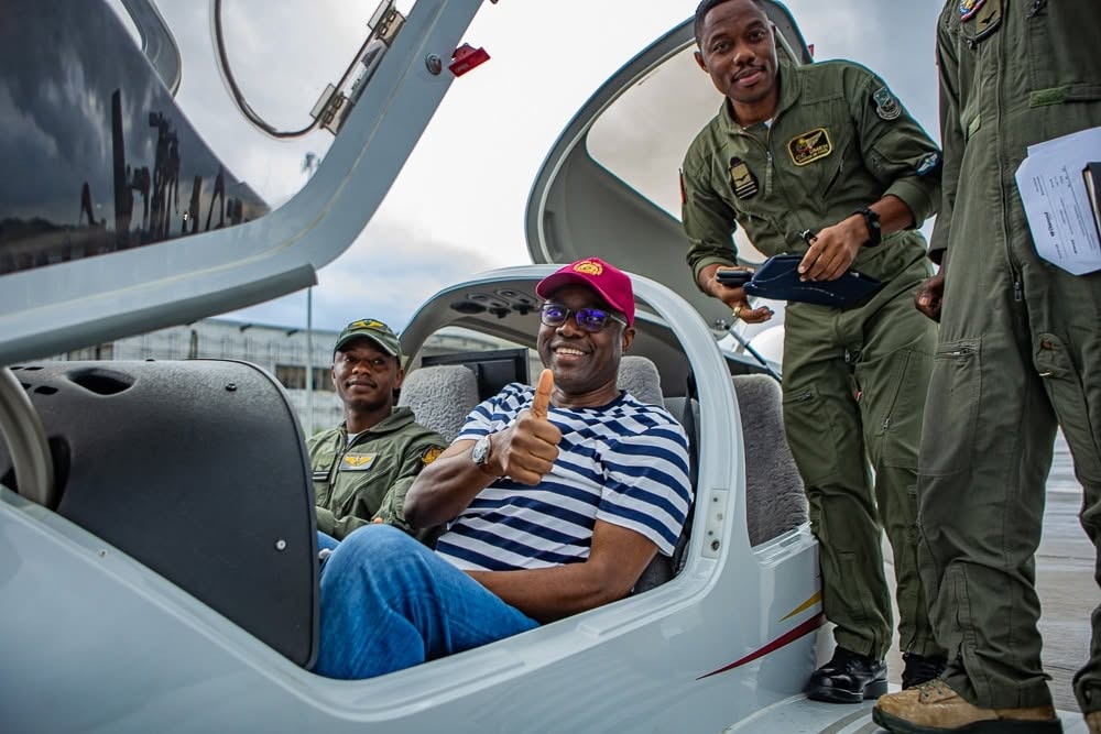Man wearing a striped shirt and red cap sits in a small airplane cockpit, giving a thumbs-up as three pilots stand nearby outside the open canopy.