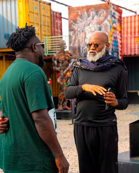 Two men chat in a colorful outdoor setting with shipping containers in the background; the older man with a white beard and sunglasses gestures while speaking.