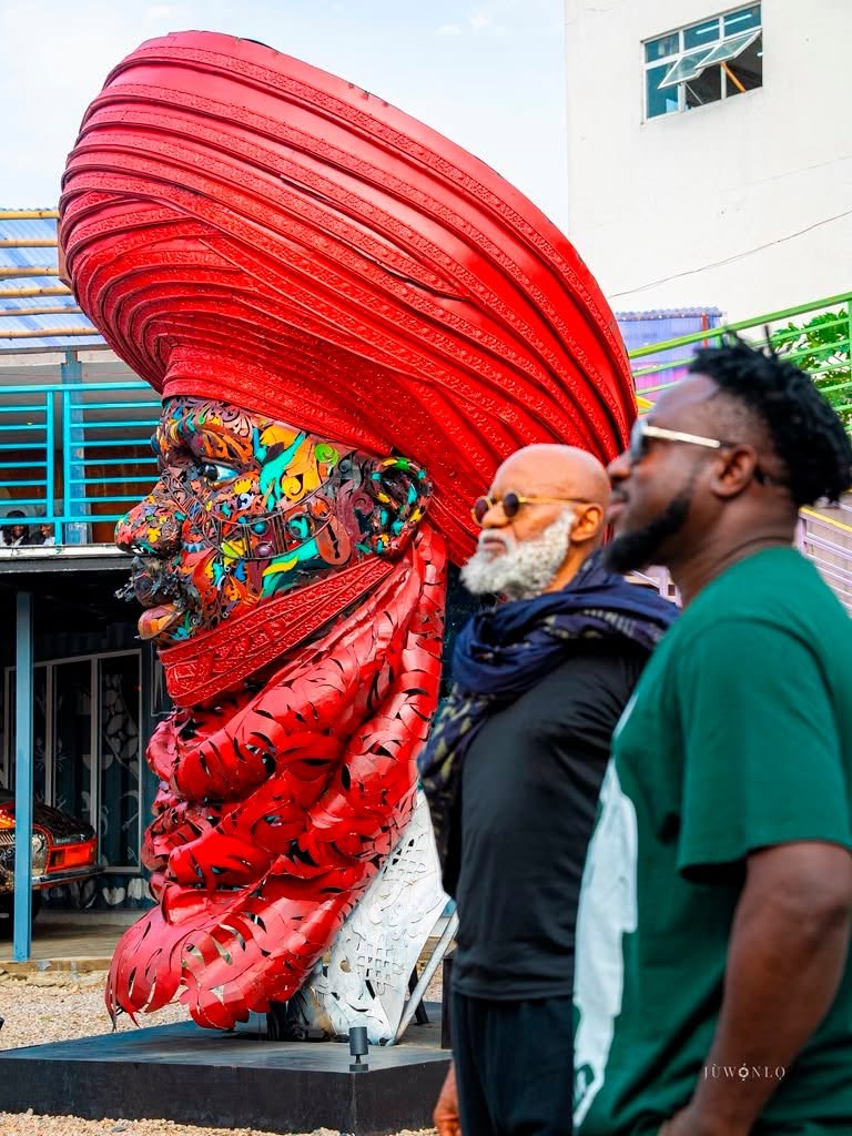 Vibrant sculpture of a head with a large red headdress and a colorful painted face, outdoors with two men nearby for scale and interest.