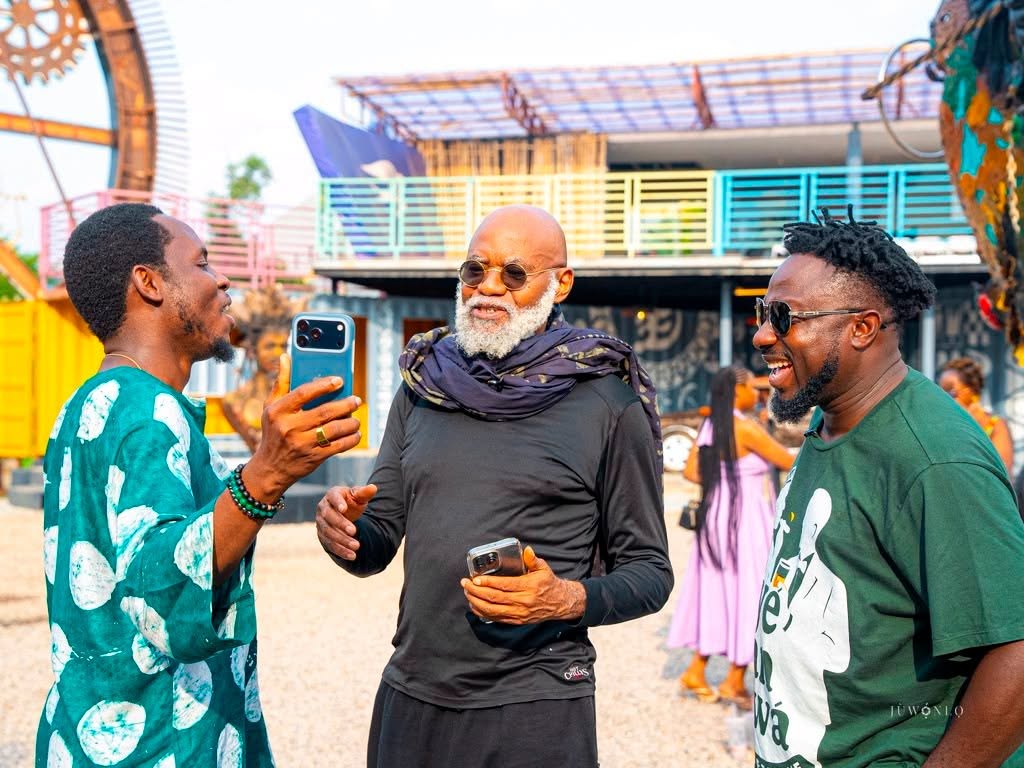 Three men chat and laugh at an outdoor festival; the man on the left holds a blue smartphone, the center man wears a scarf and sunglasses, and the man on the right wears a green graphic tee. colorful rides and people are in the background.
