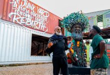 Two men stand beside a large tree-crowned sculpture made of recycled materials, near a graffiti-covered shipping container.