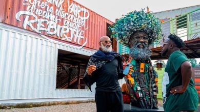 Moyo Okediji 4 - The Times of Abuja Two men stand beside a large tree-crowned sculpture made of recycled materials, near a graffiti-covered shipping container.