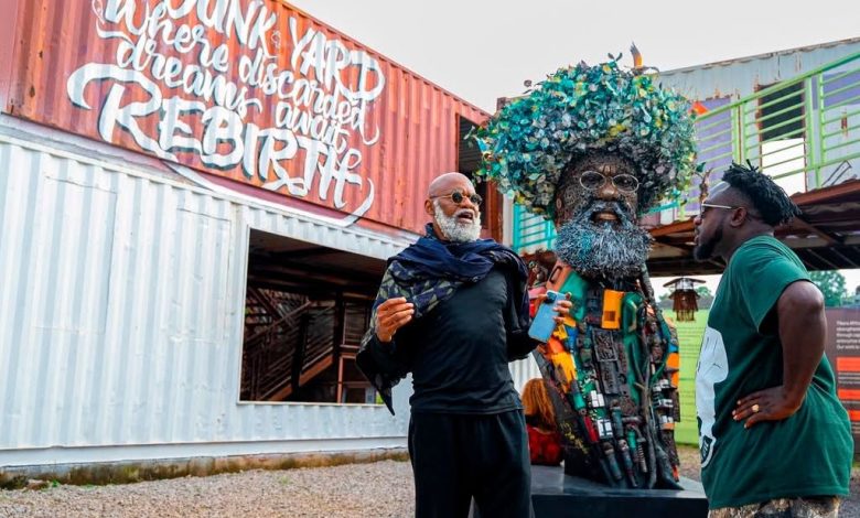 Two men stand beside a large tree-crowned sculpture made of recycled materials, near a graffiti-covered shipping container.