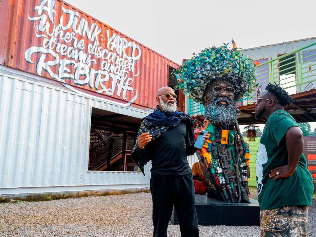 Two men stand beside a large tree-crowned sculpture made of recycled materials, near a graffiti-covered shipping container.