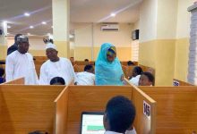 People in a busy office with wooden cubicles; a woman in a blue hijab assists a seated man while two men stand nearby in white traditional attire.