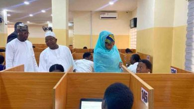 People in a busy office with wooden cubicles; a woman in a blue hijab assists a seated man while two men stand nearby in white traditional attire.