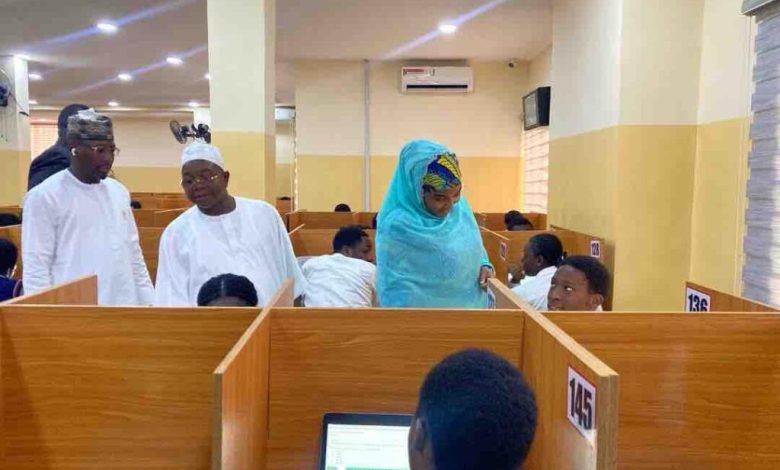 NECO CBT - The Times of Abuja People in a busy office with wooden cubicles; a woman in a blue hijab assists a seated man while two men stand nearby in white traditional attire.