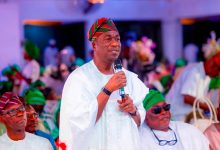 Man in white traditional attire speaks into a microphone at a festive gathering, wearing a red‑green striped cap.
