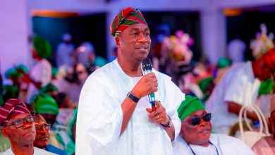 Man in white traditional attire speaks into a microphone at a festive gathering, wearing a red‑green striped cap.