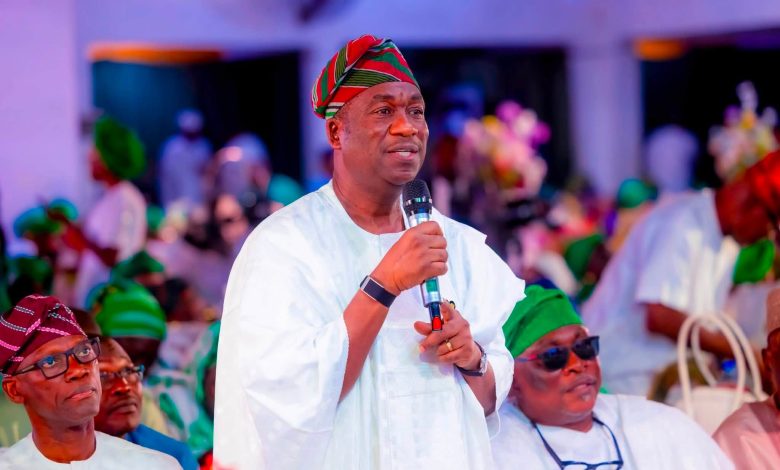 Man in white traditional attire speaks into a microphone at a festive gathering, wearing a red‑green striped cap.