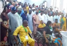 Diverse indoor ceremony with people in wheelchairs at the front and a large crowd standing behind, many wearing lanyards and colorful traditional clothing.