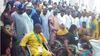 Diverse indoor ceremony with people in wheelchairs at the front and a large crowd standing behind, many wearing lanyards and colorful traditional clothing.