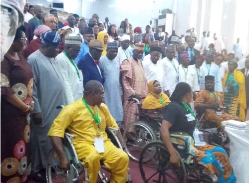 PWDs at JAMB event - The Times of Abuja Diverse indoor ceremony with people in wheelchairs at the front and a large crowd standing behind, many wearing lanyards and colorful traditional clothing.