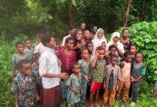 Group of African children and a few adults posing together in a lush, green outdoor setting with trees and plants nearby, wearing colorful clothing.