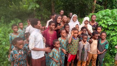 Group of African children and a few adults posing together in a lush, green outdoor setting with trees and plants nearby, wearing colorful clothing.