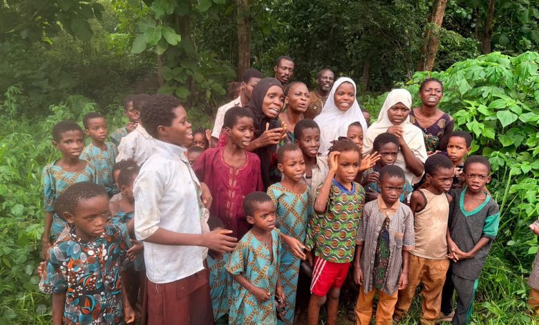 Group of African children and a few adults posing together in a lush, green outdoor setting with trees and plants nearby, wearing colorful clothing.