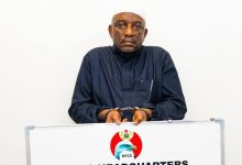 Man in a dark traditional outfit and white cap standing behind a podium with the EFCC logo, looking at the camera during a press event.