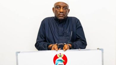 Man in a dark traditional outfit and white cap standing behind a podium with the EFCC logo, looking at the camera during a press event.