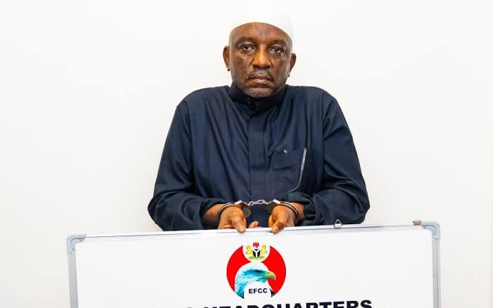 Saleh-Mamman - The Times of Abuja Man in a dark traditional outfit and white cap standing behind a podium with the EFCC logo, looking at the camera during a press event.
