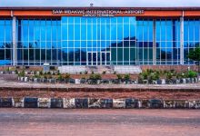 Front view of Sam Mbakwe International Airport Cargo Terminal with glass facade and a circular landscaped curb in the foreground, under a blue sky.