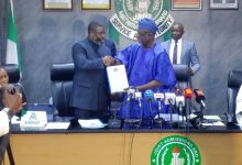 Two men shake hands across a conference table during a press briefing, one in blue traditional attire and the other in a dark suit, with a JAMB seal in the background and microphones in front.