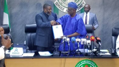 Two men shake hands across a conference table during a press briefing, one in blue traditional attire and the other in a dark suit, with a JAMB seal in the background and microphones in front.