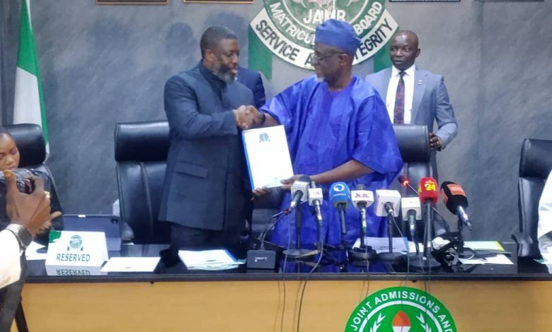 Two men shake hands across a conference table during a press briefing, one in blue traditional attire and the other in a dark suit, with a JAMB seal in the background and microphones in front.