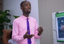 Man in a pink shirt and purple tie giving a presentation in a modern office, gesturing with his hands while holding a phone; a digital policy poster is nearby.