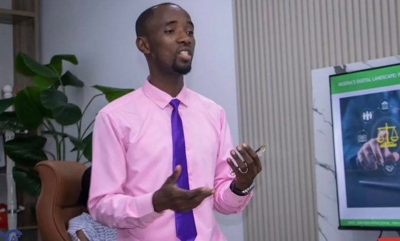 Stanley Ugagbe - The Times of Abuja Man in a pink shirt and purple tie giving a presentation in a modern office, gesturing with his hands while holding a phone; a digital policy poster is nearby.