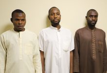 Three men stand side by side against a pale wall, wearing traditional long robes in cream, white, and brown with neutral expressions.