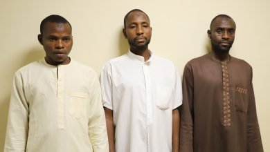 Three men stand side by side against a pale wall, wearing traditional long robes in cream, white, and brown with neutral expressions.