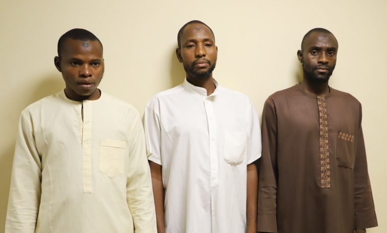 Three suspects - The Times of Abuja Three men stand side by side against a pale wall, wearing traditional long robes in cream, white, and brown with neutral expressions.